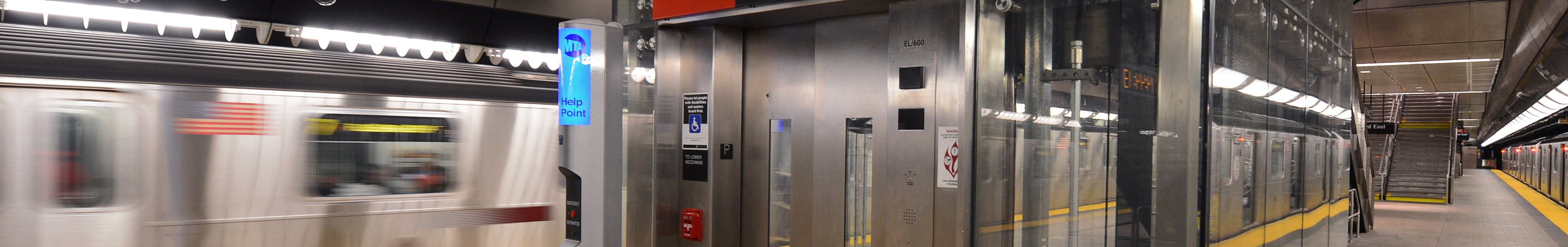 Wide photo of an elevator. On the left a train is driving by, on the right you can see the platform down to some stairs. 