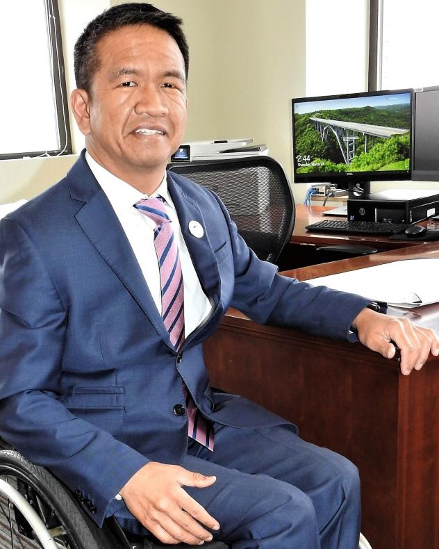 A man in a blue suit with a white shirt and a striped tie. He is using a wheelchair, and is next to a desk.