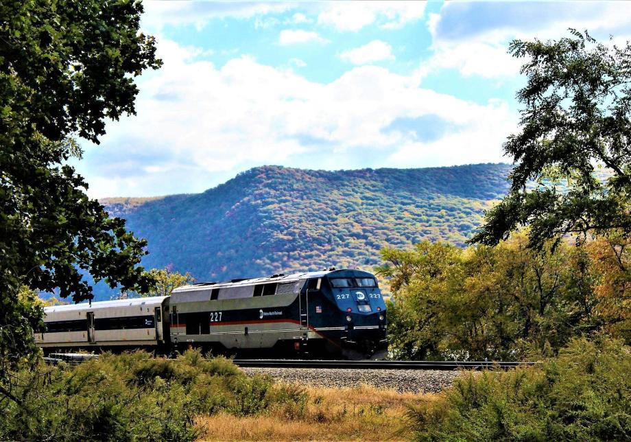 A train traveling along a track, in front of a mountain and trees with fall colors. 