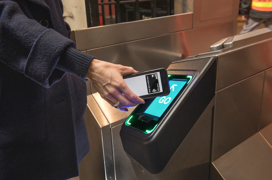 A person holds an iPhone up to a small display on a subway turnstile. The display is flashing green and saying Go.