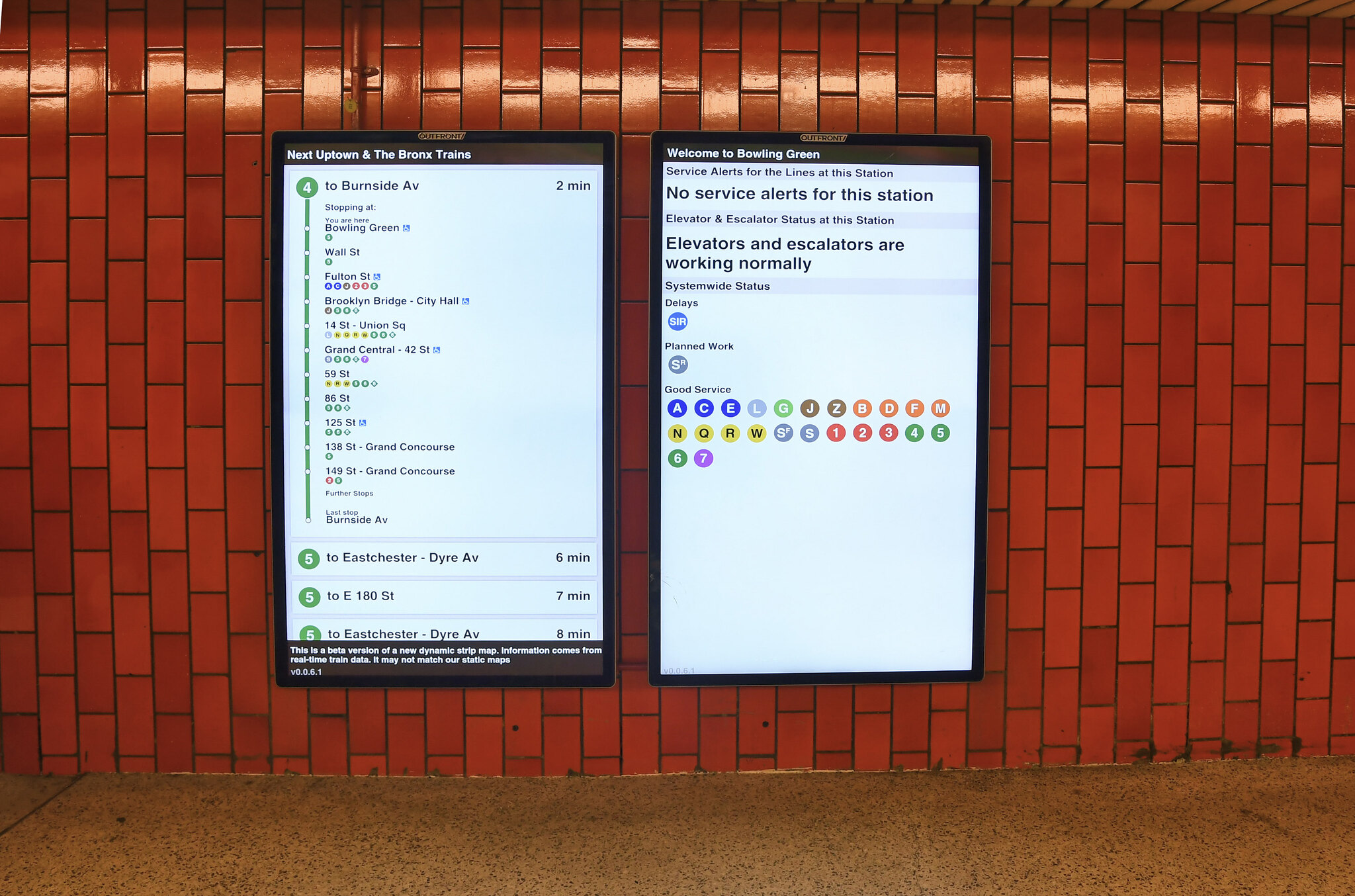 Two digital screens in the subway mounted over red tile. Screens show stop information and system status