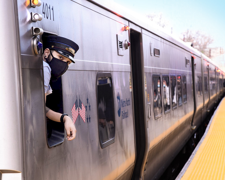 A masked Metro-North operator is leaning out the window while the train is stopped at a platform. 