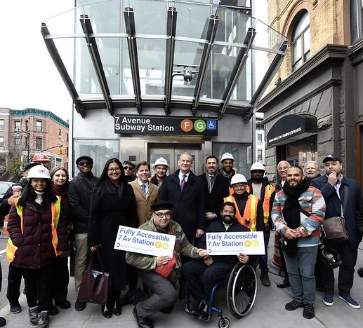 MTA Officials, construction workers, and disability advocates smile in front of street-level elevator at 7 Avenue F, G station.