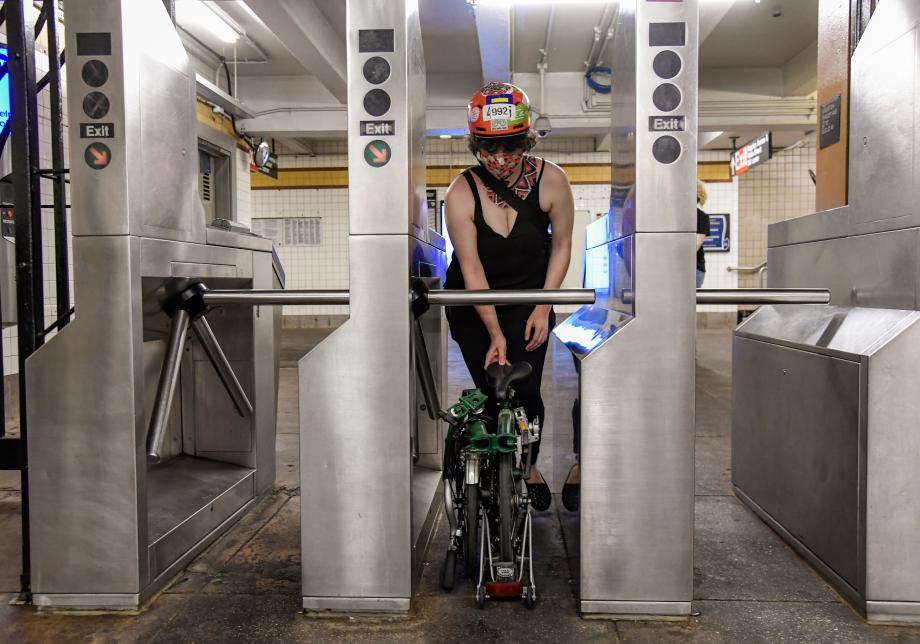 A woman rolls a green folding bike through a subway turnstile. The bike is folded up and fits underneath the turnstile.