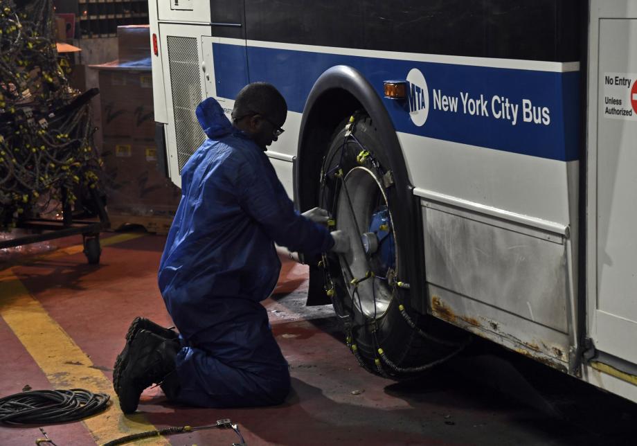 A person kneels to put snow chains on the rear tire of a bus. The words "New York City Bus" are visible on the side of the vehicle.