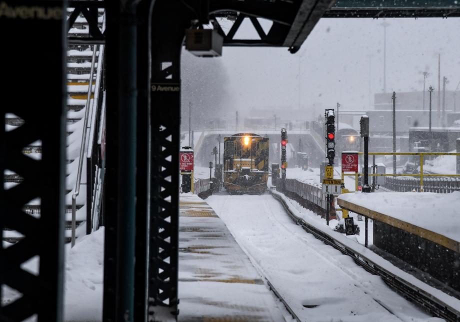A yellow and black subway train car approaches an outdoor subway station. Snow is swirling around the train.