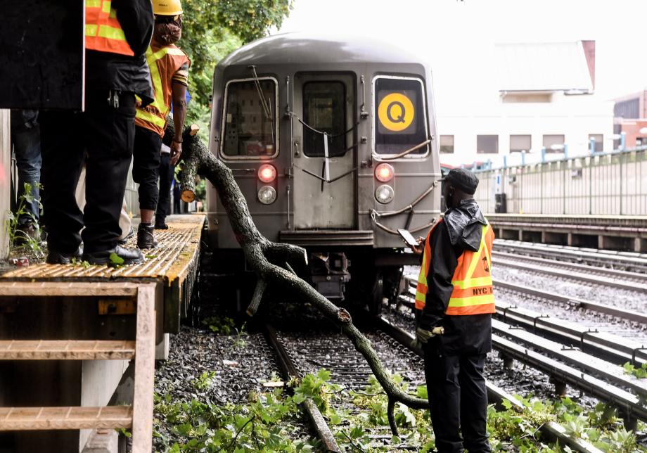 Subway workers stand near a broken tree branch that is extending onto a subway track. A subway car is stuck behind the branch. 