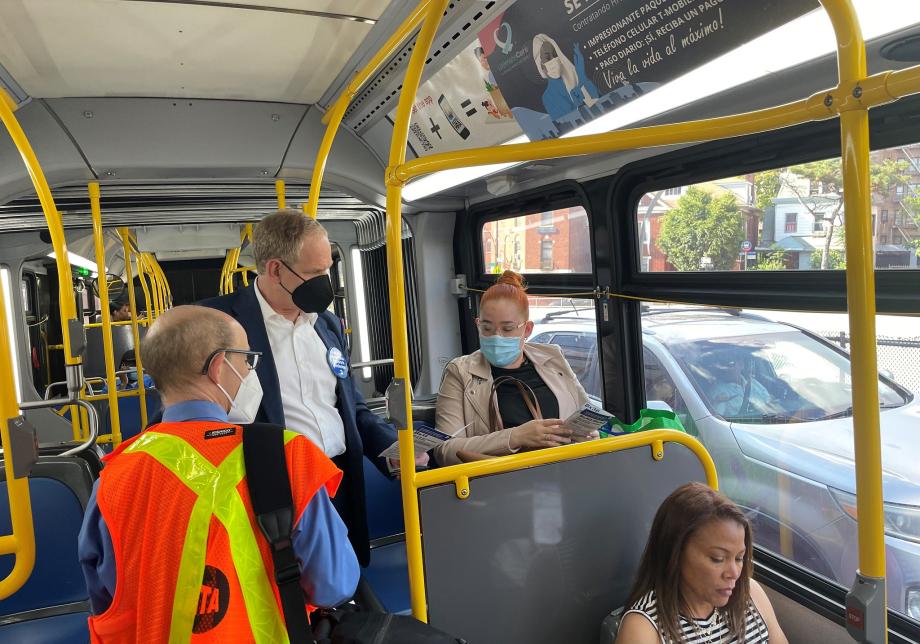 Two men are talking to a woman on a bus. One man is wearing an orange work vest, and the other is wearing a suit. The woman has red hair and is wearing a face mask.