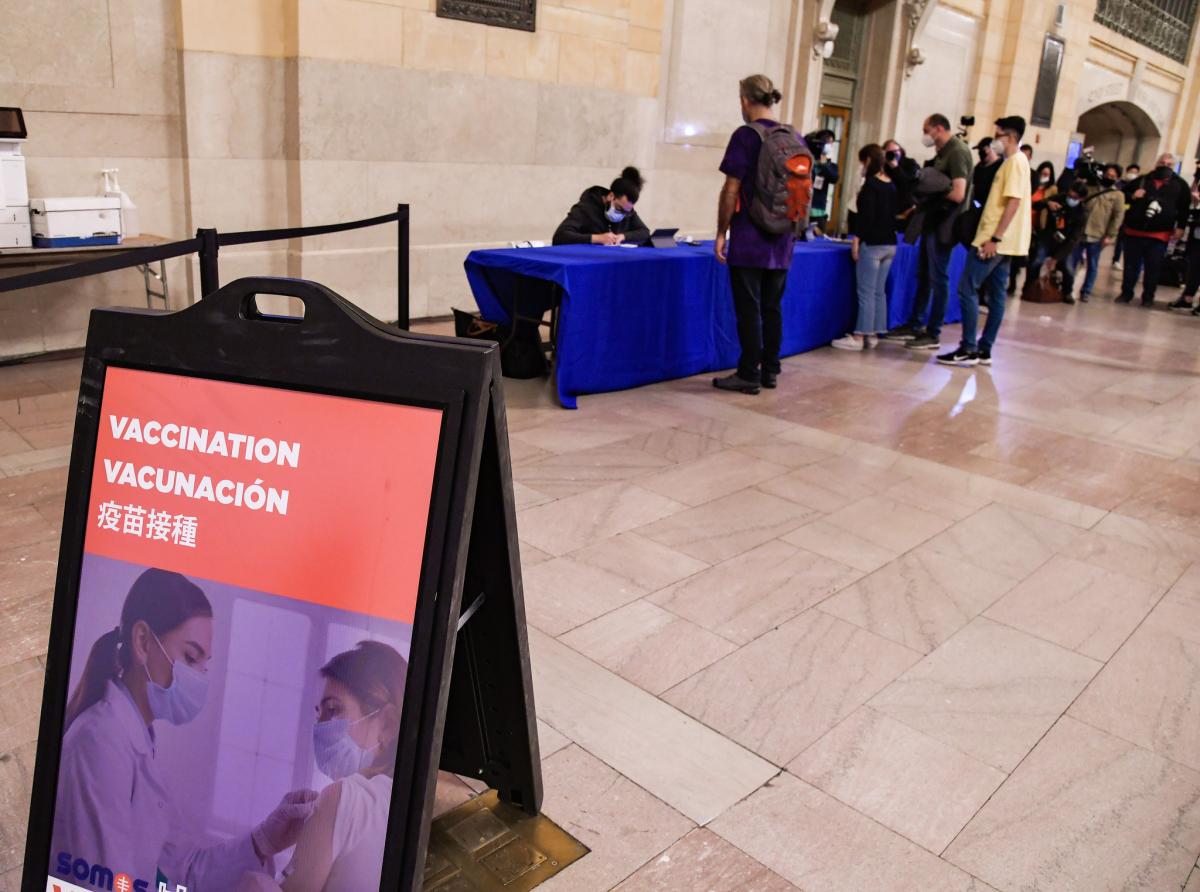 Sandwich board in foreground on limestone floor of Grand Central Terminal reading "Vaccination" in multiple languages. Blue-skirted registration table with line of people in background.