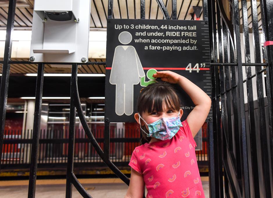 A young girl stands near a sign saying, “Up to 3 children, 44 inches tall and under, ride free when accompanied by a fare-paying adult.” She’s holding her hand near the line measuring 44 inches, which is above her head.