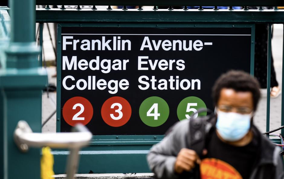 A man with a small Afro, wearing glasses, a backpack, and a face mask, exits a subway station. The sign behind him shows subway bullets for the 2, 3, 4, and 5 trains and says "Franklin Avenue-Medgar Evers College Station."
